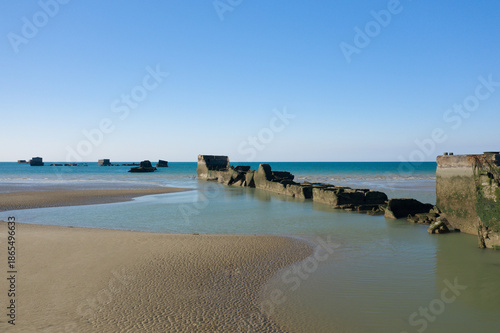 Quadro su tela Concrete ruins of a historic artificial port stretch into calm turquoise waters at low tide, with smooth sand and a clear blue sky creating a tranquil coastal scene near Asnelles, Normandy