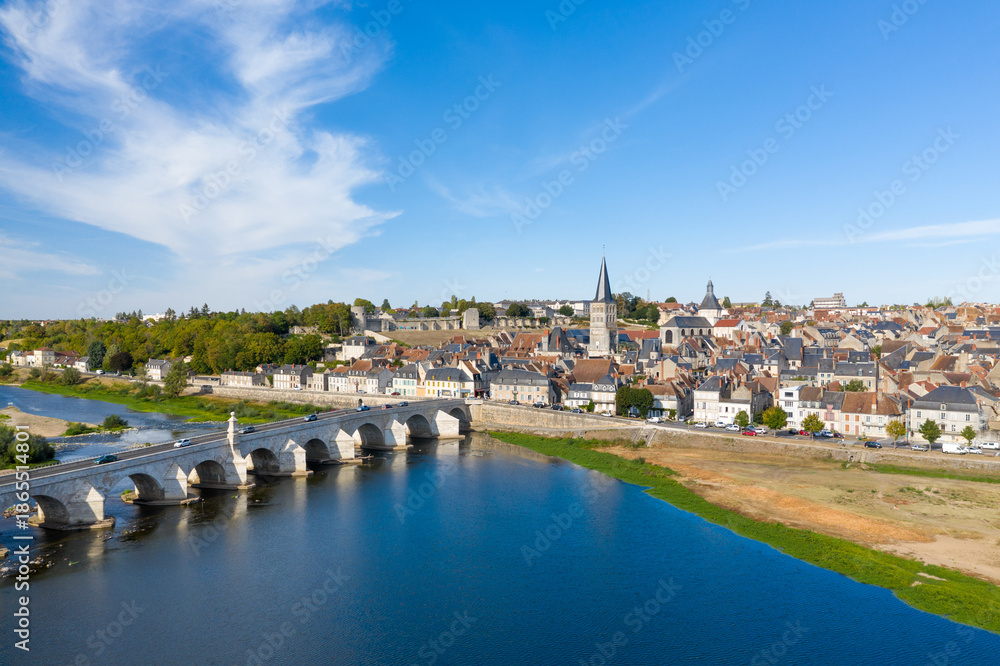 Fototapeta premium Wide view of La Charite sur Loire featuring a historic stone bridge crossing the Loire river, traditional rooftops, and a prominent church spire under a clear blue sky. The image highlights the blend