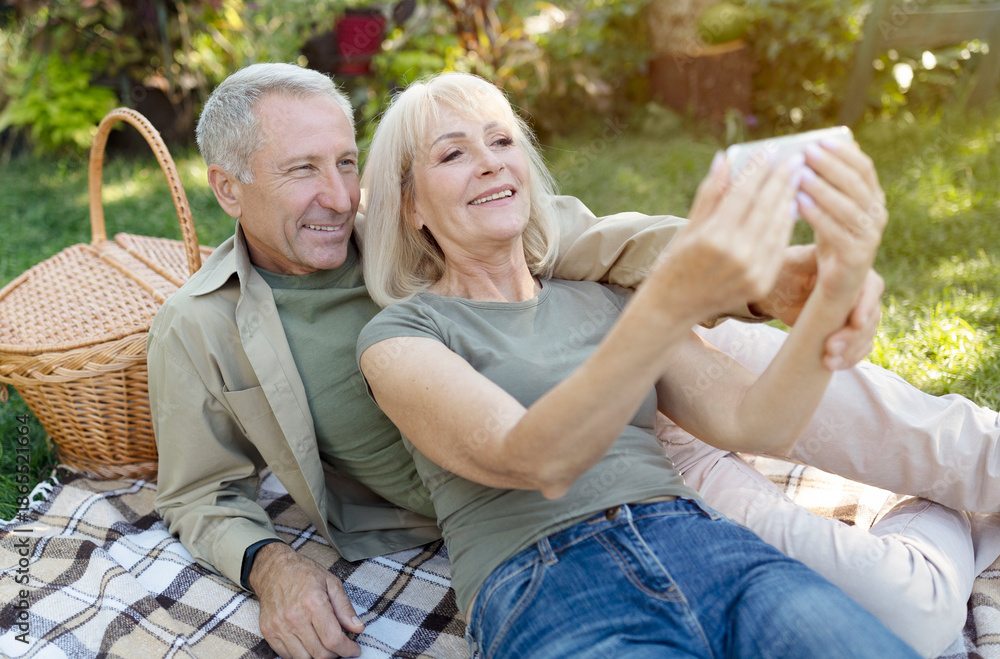 Fototapeta premium Happy senior spouses video calling on smartphone while having picnic and resting in garden at warm spring day, copy space. Elderly couple capturing happy moments, taking selfie
