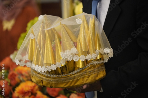 A close-up shot of a man in a black suit carrying a traditional Indonesian wedding gift (seserahan). The gift consists of a bunch of bananas individually wrapped in elegant gold cone-shaped paper, arr
