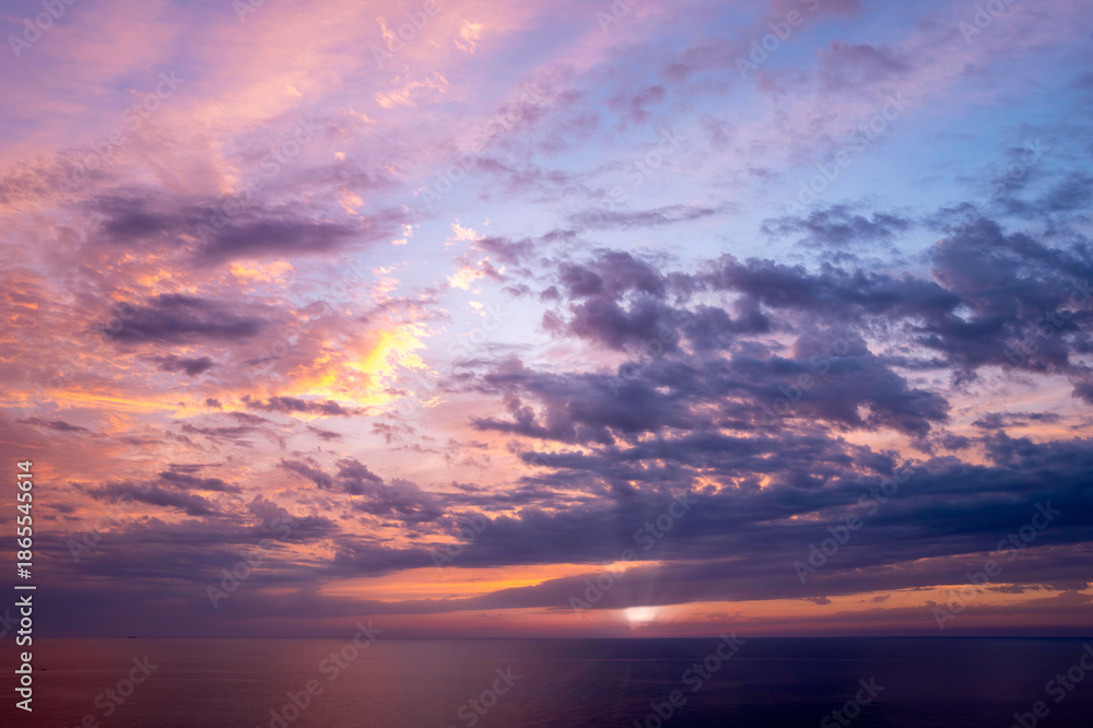 Fototapeta premium Golden and lavender clouds scatter across the sky as the sun sets above the calm Mediterranean Sea, casting soft reflections and creating a luminous, tranquil atmosphere.