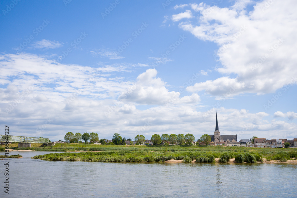Obraz premium Peaceful riverside scene with a pointed church steeple, leafy trees, and a metal bridge spanning the Loire under a bright blue sky with scattered clouds.