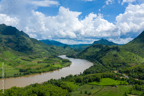 A wide river winds through lush green hills and patchwork fields beneath dramatic cumulus clouds in northern Laos. The landscape features rolling mountains, dense vegetation, and vibrant summer light.