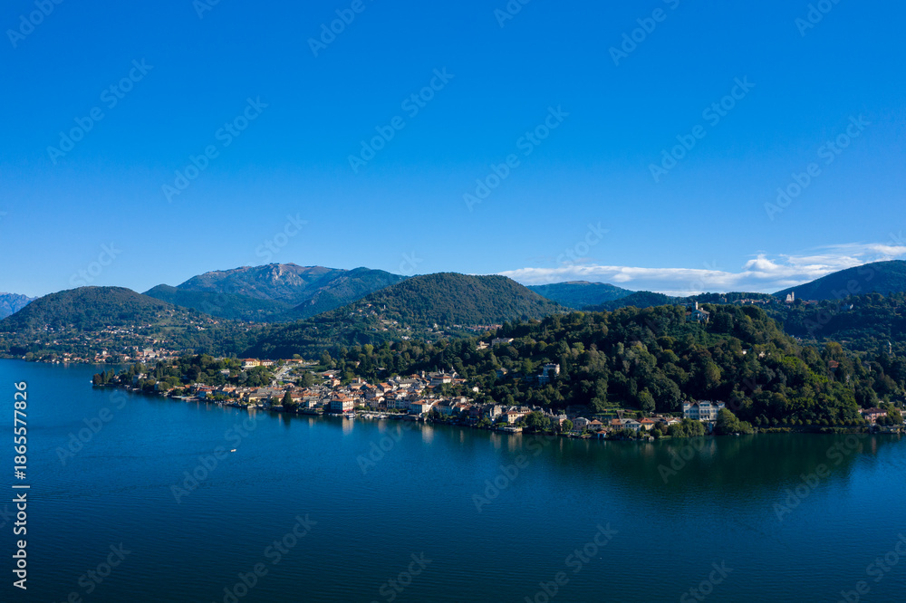 Obraz premium Sweeping view of a lakeside town nestled at the foot of densely forested hills along Lake Orta in northern Italy. The tranquil blue water reflects the surrounding greenery and distant mountain slopes
