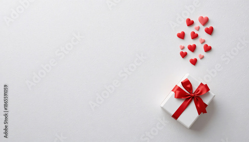 A white gift box with a red ribbon and bow surrounded by small red and pink hearts on a white