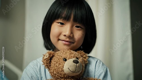 Smiling Asian girl in hospital bed holding teddy bear for comfort