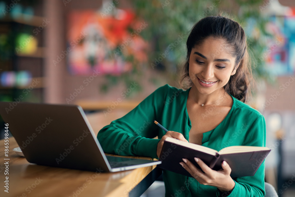 Fototapeta premium Beautiful unemployed indian lady looking for job online, sitting alone at cafe, using laptop, taking notes and smiling, getting ready for job interview, websurfing, copy space
