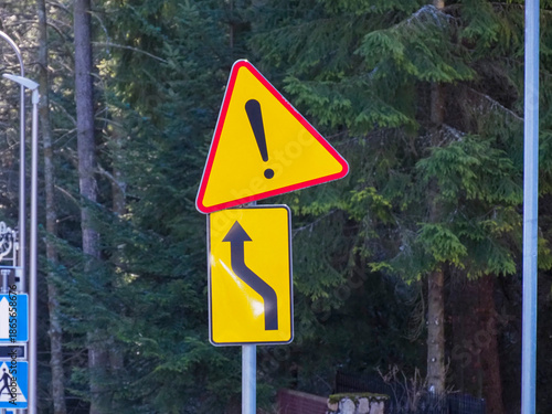 Yellow road sign with exclamation mark and winding road symbol against a forest background.