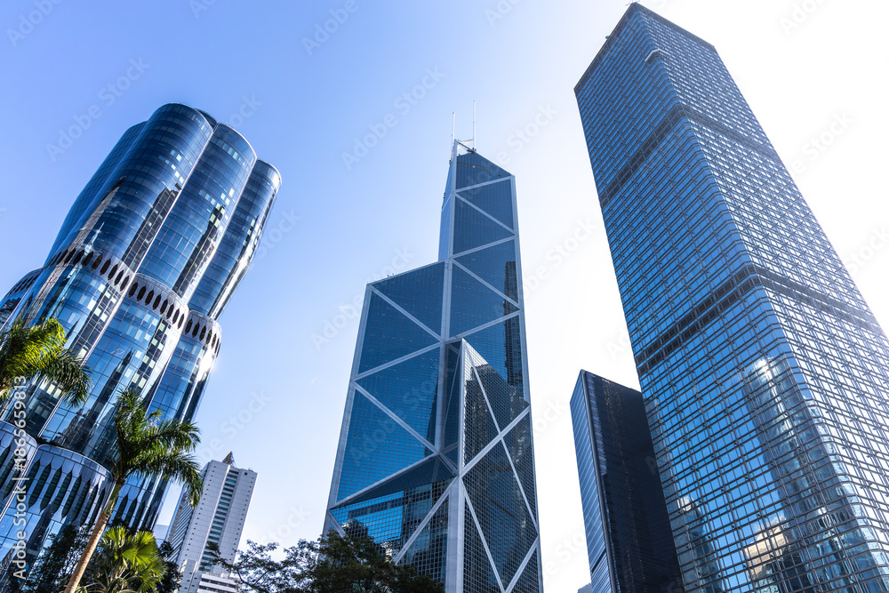 Obraz premium Modern skyscrapers in Hong Kong with glass facades and palm trees. Urban cityscape under clear blue sky.