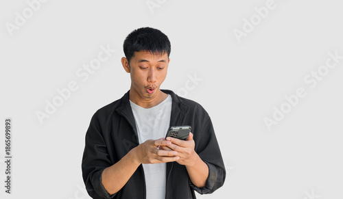 A young man playing with his smartphone makes a surprised face when he sees something exciting on his phone isolated on a grey background. The concept is related to social media usage.