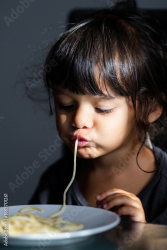 Funny messy face with spaghetti. Toddler eating noodles. Cute childhood emotion during eating. Kid eating spaghetti meal at home. Messy spaghetti mouth and lips moment. Toddler eating pasta indoors.
