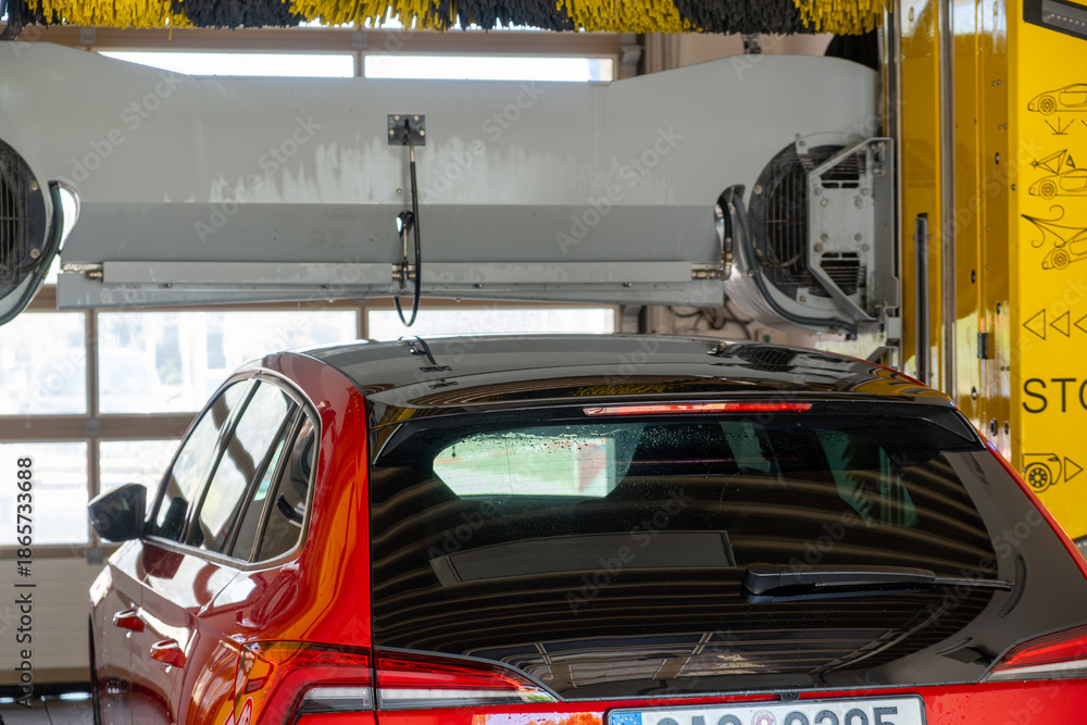 Fototapeta premium Red car passing through an automatic car wash, water and brushes cleaning the vehicle.