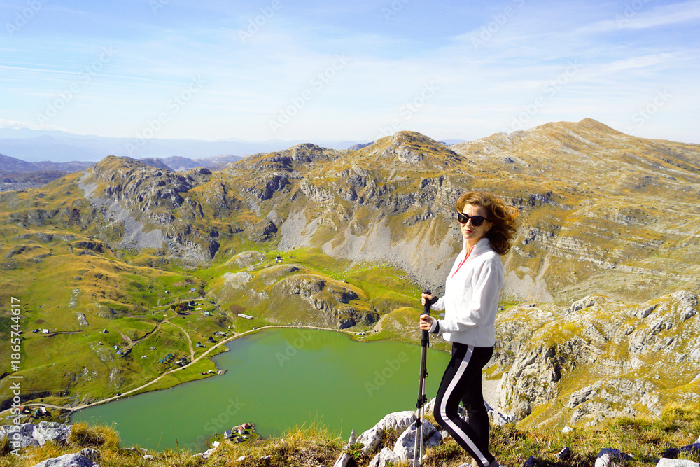 Naklejka premium Woman with trekking poles near Kapetanovo Lake in Moraca mountains. Symbolizes wellness through movement, solo adventure travel, balance between effort and calm environment, and nature focused tourism