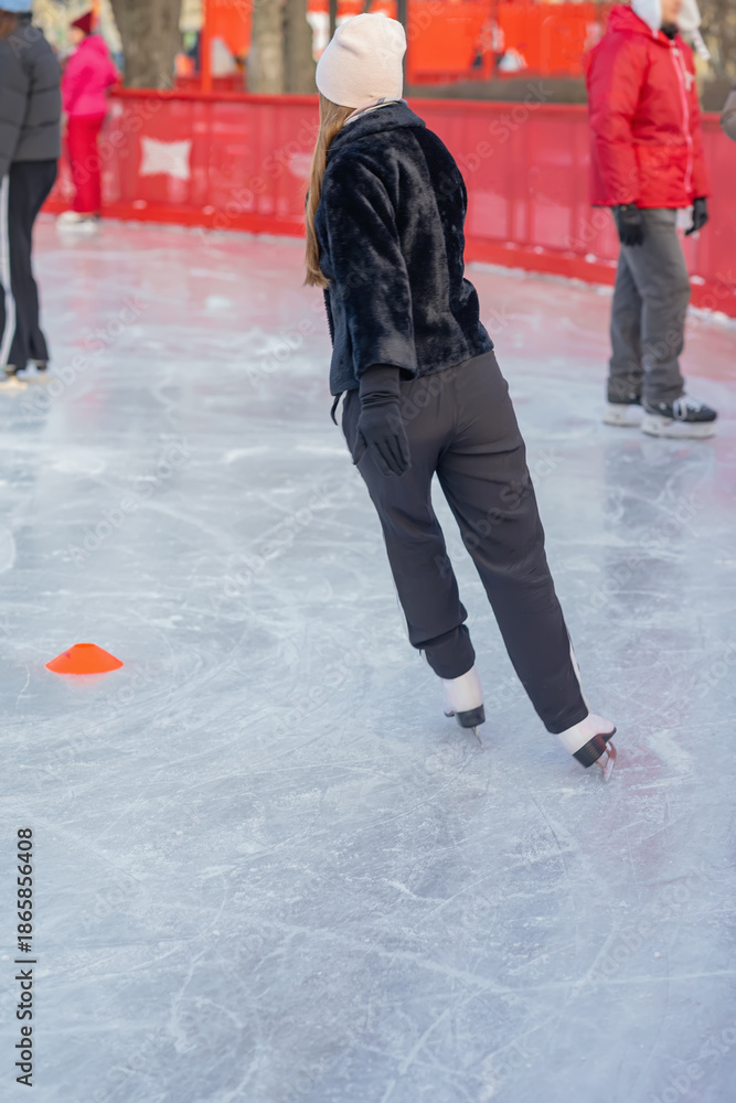 Obraz premium Rear view of woman on ice in black jacket. Woman skating on outdoor ice rink in winter clothes. Concept of sport, winter fun and seasonal recreation