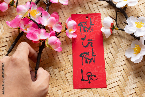 A hand holding a pink peach blossom branch next to a prosperity tag, representing preparations for a wealthy and successful Vietnamese Tet