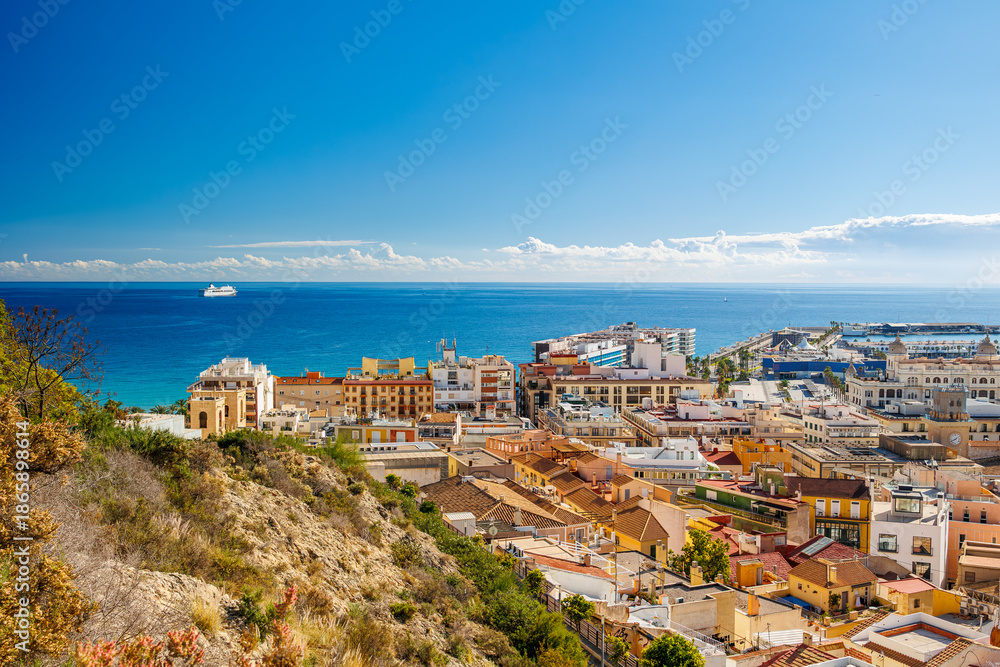 Fototapeta premium Coastal city panorama with sea and harbor view, Panoramic view of a Mediterranean coastal city with colorful rooftops, harbor, hillside foreground, and deep blue sea under clear sky.