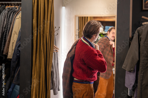 Male customer in men's wear second hand boutique trying on vintage fashionable checkered blazer in front of mirror in open fitting room. Mustachioed elegant man in jumper fits on jacket in shop.