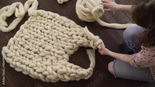 A young woman weaves a product made of thick merino wool with her hands while sitting on the floor in her apartment. Close-up of hands. Small business on maternity leave.