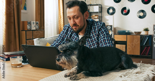 A focused young man with a disability sits in a wheelchair, working online from home in casual clothes, smiling as his favourite puppy dog stays nearby, offering calm support.