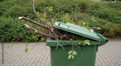 An overflowing green yard waste trash bin with branches and leaves outdoors on a paved surface.