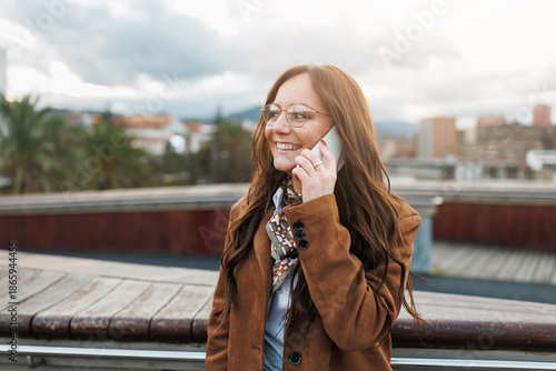 Cheerful woman with long hair and glasses talking on smartphone, wearing brown coat, standing outside in city with cloudy sky and soft light
