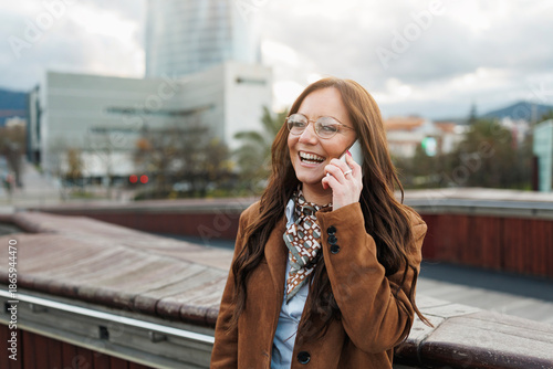 Woman wearing brown coat and glasses laughing while talking on phone, urban background with modern buildings, concept of communication
