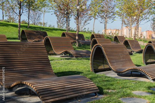 Modern curved wooden benches are on green lawn surrounded by trees on gentle slope against backdrop of city buildings. Recreation area is on hill in Oblaka Park, Krasnodar City Park or Galitsky Park.