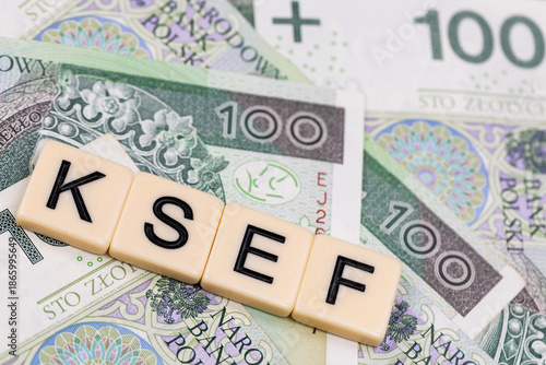 Blocks with the abbreviation KSEF, standing for National E-invoice System, are placed against a background of loosely spread Polish 100 PLN banknotes. The photo was taken under artificial lighting.