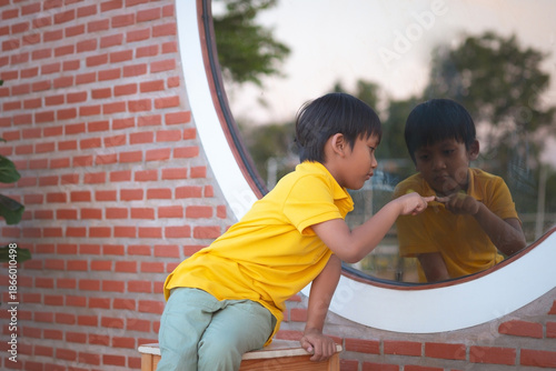A boy is sitting and pointing his finger at a mirror which reflects his image, feeling lonely.