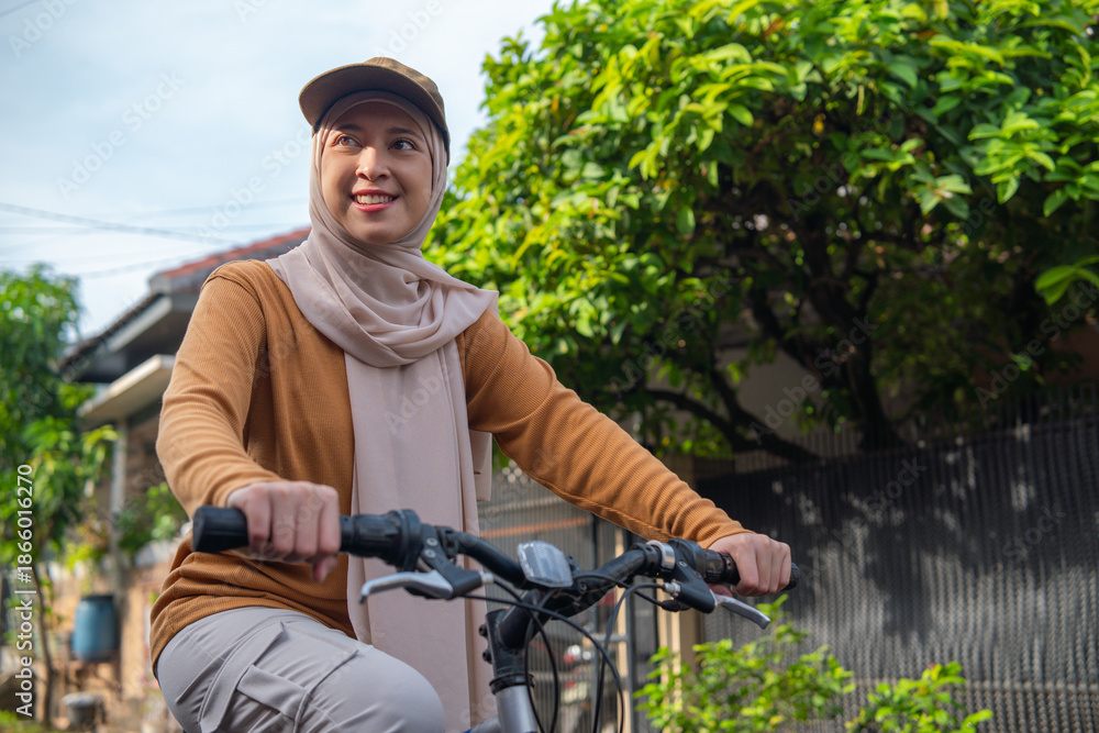 Obraz premium Asian Muslim woman wearing a hijab rides a bicycle along a tree-lined street, enjoying a sunny day.