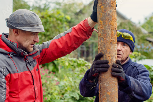 Fotografi Two focused men in workwear, wearing gloves, collaboratively handling a rusty metal pipe outdoors