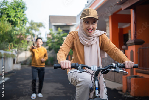 Wallpaper Mural A smiling woman wearing a hijab and cap rides a bicycle down a sunny residential street while a friend waves in the background Torontodigital.ca