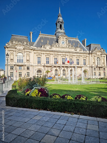 The Hotel de Ville in Tours, France. Grand Neoclassical building with ornate facade and classic clock tower, decorated with beautiful tiered fountain and lush green garden with vibrant flower beds