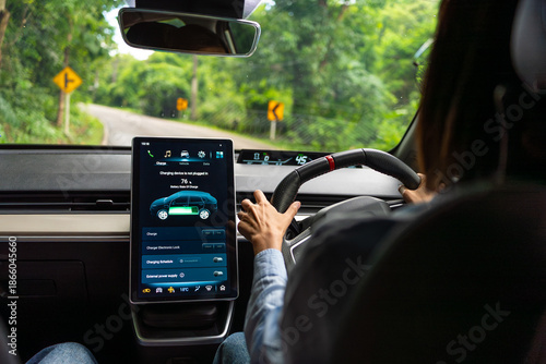 Asian woman driver electric car along a scenic winding road, surrounded by lush greenery. The digital touchscreen displays the electric car battery level and charging options. Future energy concept.