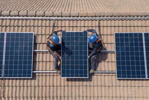 Professional technician wearing safety harness and lifeline installing solar panels on high rise building rooftop with urban city skyline view for renewable green energy maintenance service task