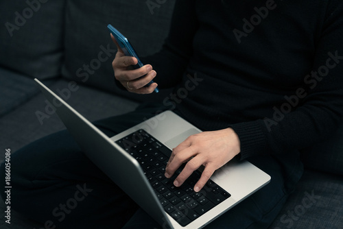 High angle cropped view of woman sitting on grey sofa working on laptop and checking smartphone simultaneously representing remote work and digital multitasking efficiency in modern home lifestyle