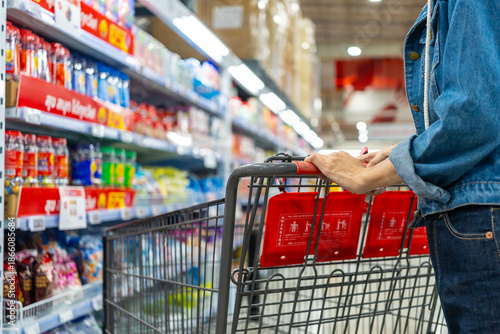 Image of asian woman shopper pushing a red shopping cart in a warehouse store aisle, surrounded by shelves full of bottled water and other items ready for customers to add to their carts.