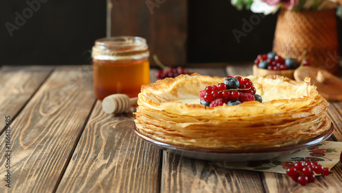 Delicious stack of thin pancakes served with fresh berries and honey on wooden table with flowers in background