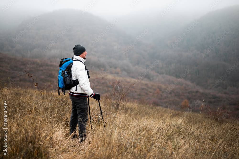 Fototapeta premium Happy hiker in warm clothing standing in nature with backpack and hiking poles, enjoying a peaceful moment in winter wilderness.