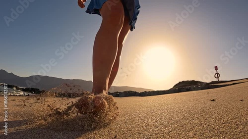 LOW ANGLE VIEW, SLOW MOTION, LENS FLARE: Young lady in blue skort during a sunset walk along sandy shore. She is walking barefoot and enjoys carefree summer moments on holidays at Canary Islands.