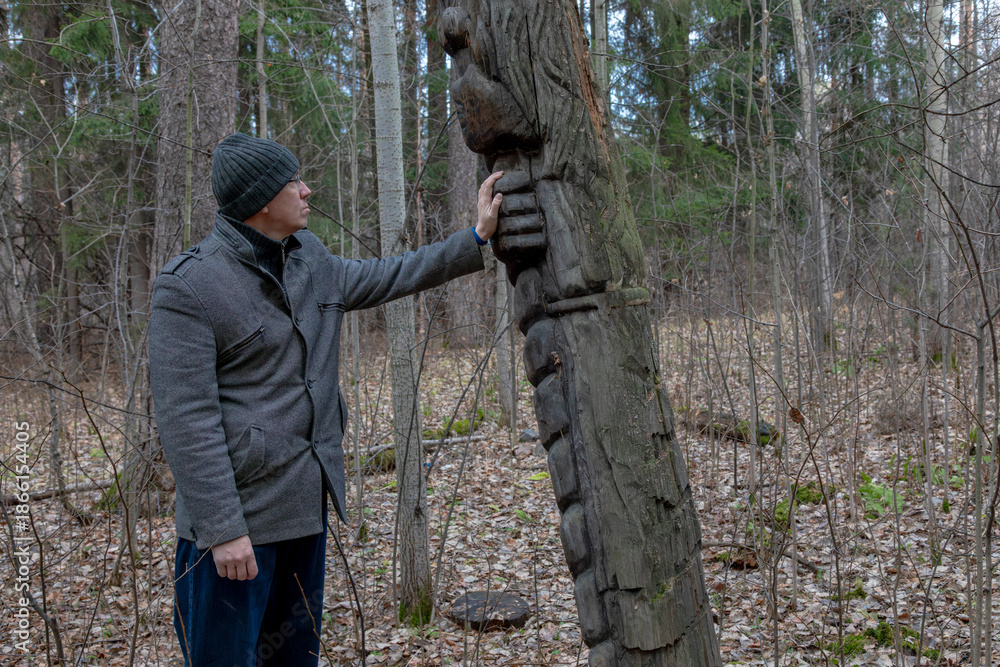 Naklejka premium Adult man in a gray coat touches an ancient wooden idol in an autumn forest. Mysterious pagan altar or sculpture in a wild woodland.