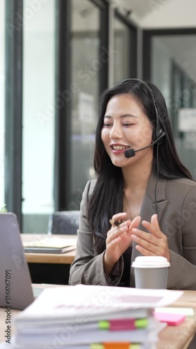Professional Asian woman wearing a headset and working on a laptop while providing customer support in a modern corporate office environment.
