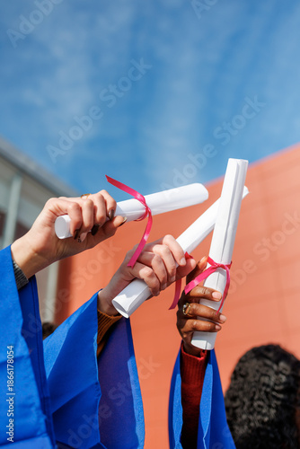 Diverse students proudly raising their rolled diplomas with pink ribbons towards a blue sky