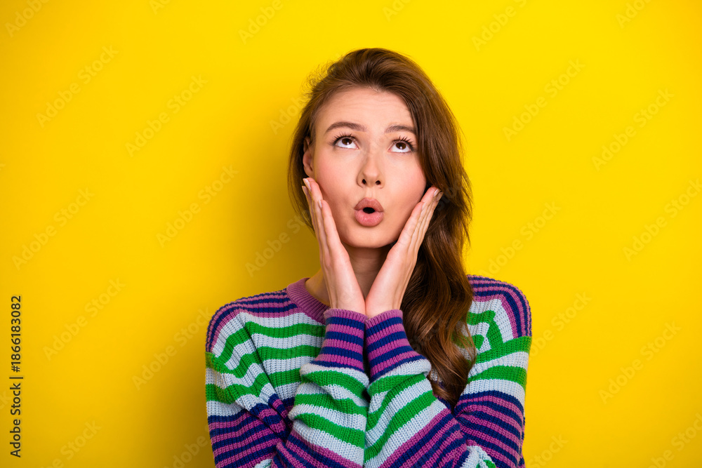 Fototapeta premium Young woman in colorful striped sweater looks surprised with hands on cheeks yellow background smiling and conveying playful casual fashion mood