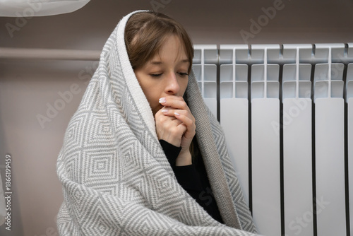 Girl wrapped in a warm blanket sitting by a white radiator, rubbing her hands and trying to stay warm indoors during cold winter heating worries and energy cost concerns