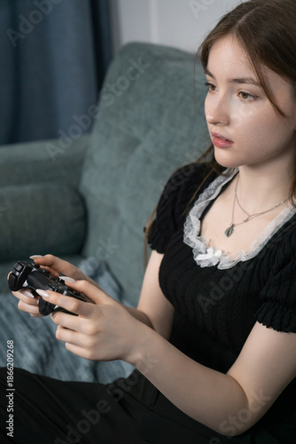 Young girl holding a joystick controller and playing video games sitting on a comfortable sofa in her living room, enjoying her leisure time. Vertical photo