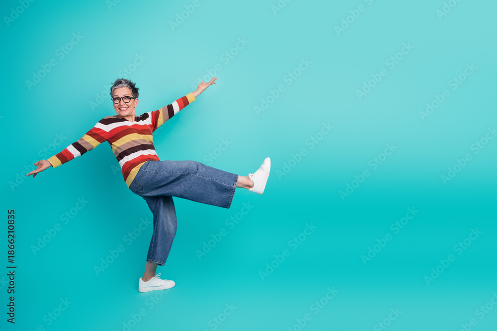 Obraz premium Aged mature woman with short hair wear sweater jeans sneakers smiles while kicking one leg dynamic pose blue background lifestyle advertising fashion