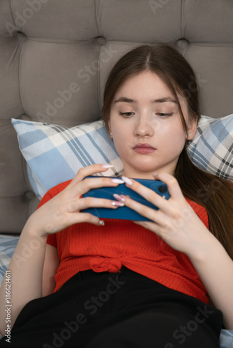 Comfortable young girl lounging on a sofa, engrossed in her smartphone, enjoying the convenience of mobile technology while relaxing in a cozy indoor setting. Vertical photo