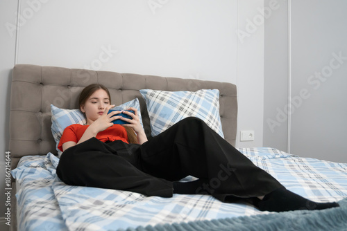 Girl lying on a comfortable bed, using her smartphone for entertainment, communication, or social media, enjoying her leisure time in a cozy home environment