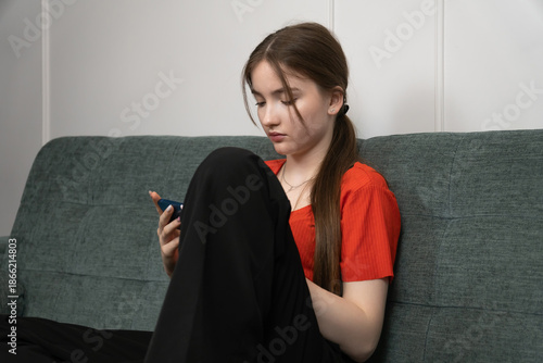 Teenager girl is playing mobile game or using social media app on blue smartphone, sitting on grey sofa at home, wearing red t-shirt and black pants